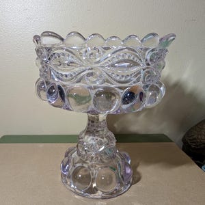 May include: A clear glass pedestal bowl featuring a scalloped rim and a repeating pattern of raised circular dots around the body. The base is also decorated with a row of raised circular dots and a central embossed floral design.