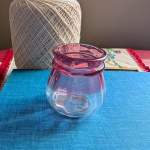 May include: A small, clear glass jar with a pink rim and base. The jar has a rounded shape and sits on a blue surface. A large spool of white thread and greenery are in the background.