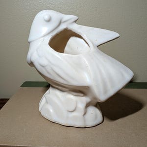 May include: A white ceramic planter shaped like a bird perched on a rock. The bird has a smooth, glossy finish with molded wing and tail feather details. The opening for the plant is located on the back of the bird, creating a functional vessel.