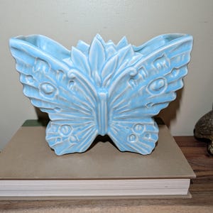 May include: A light blue ceramic planter shaped like a butterfly with raised wing details and a central leaf-like motif. The piece features a glossy finish and an open top for holding items. A ruler at the bottom shows measurements in inches.