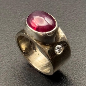 May include: A silver ring with a large, oval, deep red gemstone. A small, round, clear stone is set into the band. The ring has a thick, textured band and a bezel setting for the main stone.