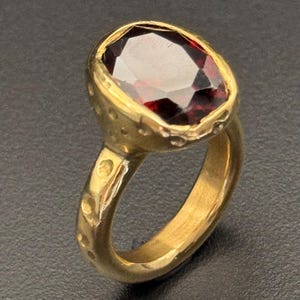 Könnte beinhalten: Ein goldener Ring mit einem großen, facettierten, ovalförmigen, tiefroten Edelstein. Der Ring hat ein strukturiertes Band mit kleinen, dekorativen Vertiefungen. Die Fassung um den Edelstein ist ebenfalls strukturiert.