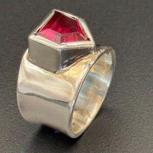 Peut inclure: Une bague en argent avec une grosse pierre pr&eacute;cieuse rouge facett&eacute;e. La pierre est sertie dans une lunette carr&eacute;e, et le corps de la bague est large et poli. La bague est pr&eacute;sent&eacute;e sur fond sombre.