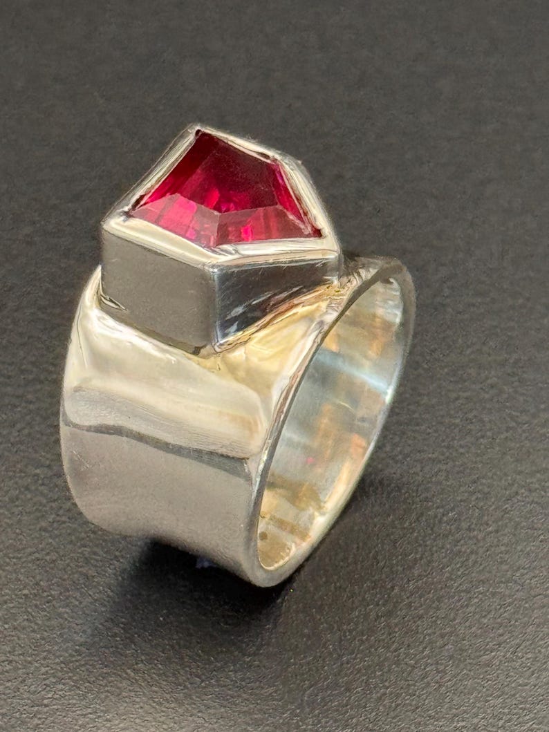 Peut inclure: Une bague en argent avec une grosse pierre pr&eacute;cieuse rouge facett&eacute;e. La pierre est sertie dans une lunette carr&eacute;e, et le corps de la bague est large et poli. La bague est pr&eacute;sent&eacute;e sur fond sombre.