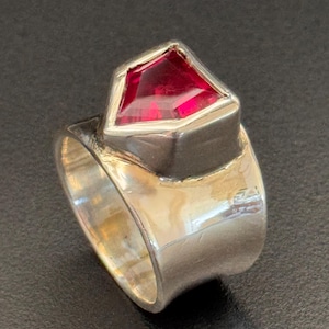 Könnte beinhalten: Ein silberner Ring mit einem großen, facettierten, roten Edelstein. Der Edelstein ist in einer geometrischen Lünette gefasst. Der Ring hat ein breites, poliertes Band. Der Ring wird vor dunklem Hintergrund gezeigt.