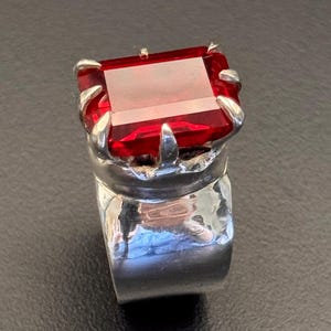 Könnte beinhalten: Ein silberner Ring mit einem großen, rechteckigen, roten Edelstein. Der Edelstein ist in einer krallenartigen Fassung gefasst, und das Band ist breit und poliert. Der Ring ist ein Statement-Schmuckstück, geeignet für besondere Anlässe.