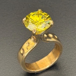 May include: A gold ring featuring a large, round, bright yellow gemstone. The band is gold with decorative accents. The ring is set against a dark background, highlighting the vibrant color of the stone.