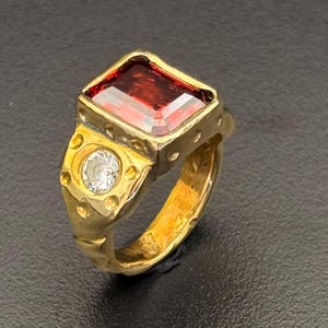 Puede incluir: Un anillo dorado con una gran gema rectangular de color rojo oscuro. El anillo también presenta una piedra más pequeña, redonda y transparente y detalles decorativos. El anillo está sobre un fondo oscuro.