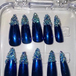 May include: A set of ten long, glossy, blue press-on nails. Each nail is adorned with a cluster of iridescent rhinestones at the cuticle. The nails are displayed in a clear plastic case.