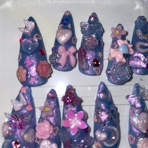 May include: A set of ten long, pointed, artificial nails. Each nail is painted a shimmering blue with purple accents and adorned with various embellishments, including hearts, flowers, bows, and glitter. The nails are arranged in a clear plastic container.