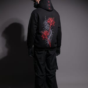 Puede incluir: Sudadera con capucha negra con un diseño floral rojo y gris en la espalda. La capucha presenta tres marcas 'X' rojas. La persona lleva pantalones, guantes y botas negros. También se usa una mascarilla negra.