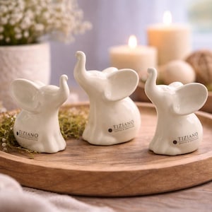 May include: Three white ceramic elephant figurines with raised trunks and large ears are arranged on a wooden tray. The figurines have the word "TIZIANO" printed on them. Candles and decorative items are in the background.