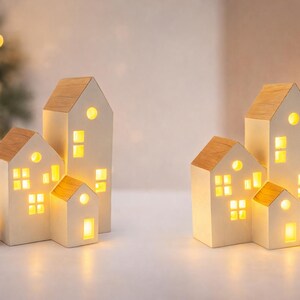 Könnte beinhalten: Set aus vier weißen Keramik-Kerzenhaltern in Hausform mit Holzdächern. Die Häuser haben beleuchtete Fenster, die ein warmes Leuchten erzeugen. Eine Kiefernzapfen und Grünzeug deuten auf ein weihnachtliches Thema hin.
