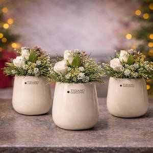 Könnte beinhalten: Drei cremefarbene Keramikvasen, jede gefüllt mit weißen Rosen und Grünzeug. Die Vasen haben eine abgerundete Form und eine glänzende Oberfläche, mit dem Logo "TIZIANO" auf der Vorderseite. Der Hintergrund zeigt verschwommene Weihnachtsbäume.