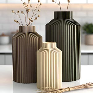 May include: Three decorative vases in varying shades of brown and beige, with vertical ribbed detailing. Each vase holds dried floral stems. A bundle of dried wheat is in the foreground. The vases are on a white surface.