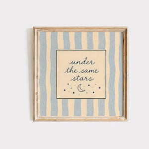 May include: Square framed art print with a light beige background and vertical blue stripes. The center features a square with the words "under the same stars" and a crescent moon and stars.