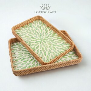 May include: Two rectangular woven rattan trays with a light brown natural finish. The interior base of each tray features a white surface decorated with a repeating green leaf-like petal pattern radiating from the centre. The brand name LOTUSCRAFT is visible.