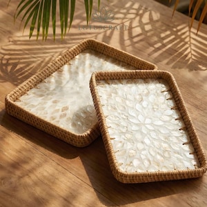 May include: Two rectangular trays featuring woven light brown rattan frames and bases inlaid with iridescent white shell pieces arranged in a repeating petal-like pattern. The trays have raised edges and a natural finish, showcasing the texture of the shell.