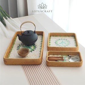 May include: A set of three woven rattan trays with light brown frames and inlaid mosaic bases featuring green and white floral patterns. The largest tray holds a black cast iron teapot and a small cup, while the smallest tray contains two metal forks.