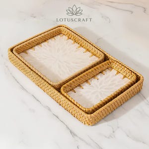 May include: Two rectangular woven rattan trays with light tan natural fibres and white resin inserts featuring a floral petal pattern. The Lotuscraft logo is visible at the top. The trays are nested together, showcasing a structured woven border.