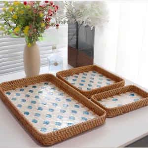 May include: Three rectangular woven rattan trays with a light brown finish. Each tray features a flat base inlaid with a white and blue scalloped shell pattern. The trays are arranged in a descending size order from left to right on a surface.