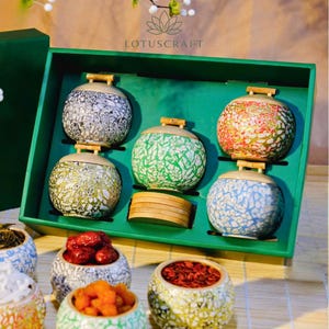 May include: A set of six decorative ceramic jars with bamboo lids, presented in a green box. The jars feature intricate patterns in various colors, including white, green, red, and blue. The brand name "LOTUSCRAFT" is visible.