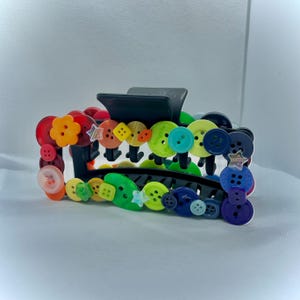 May include: A black hair claw clip decorated with an assortment of colorful buttons. The buttons are in various sizes, shapes, and colors, including red, orange, yellow, green, blue, and purple. Some buttons have star-shaped accents.