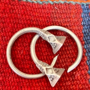 May include: Two silver bracelets with geometric designs are interlocked, set against a red woven background. The bracelets have a circular band with decorative triangular ends. The background is a vibrant red with blue and orange accents.