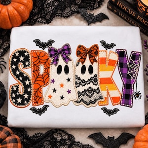 以下が含まれることがあります： 白いTシャツに、大きくカラフルな柄入りの文字で「SPOOKY」と書かれています。Sは星柄の黒、Pは蜘蛛の巣柄のオレンジ、Oはリボンをつけたお化け、Kはオレンジと白のストライプ、Yは紫のチェック柄です。