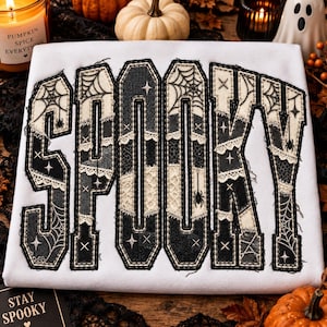 以下が含まれることがあります： 大きなブロック体の文字で「SPOOKY」と書かれた白いスウェットシャツ。文字は黒と白のパッチワーク柄で埋め尽くされており、クモの巣のデザイン、レースの縁取り、小さな星のアクセント、ぶら下がっているクモのグラフィックが施されています。