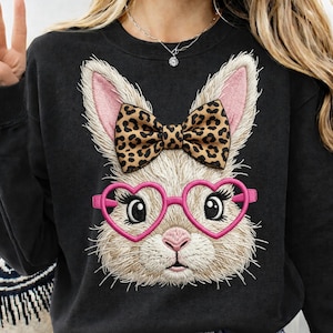 May include: Black sweatshirt featuring an embroidered bunny face with pink heart-shaped glasses and a leopard print bow. The bunny has fluffy white fur and pink inner ears. A fun and stylish design for casual wear.