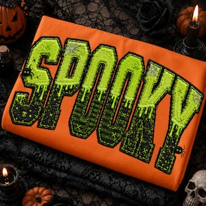 Op de afbeelding: Een oranje sweatshirt met het woord SPOOKY in grote, textuurrijke, limoengroene en zwarte letters in varsity-stijl. De letters hebben een druipend effect, spinnenwebaccenten en kleine spinnengrafieken, met een zwarte gestikte omlijning.