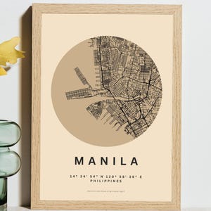 Puede incluir: Una impresión enmarcada que presenta un mapa de Manila, Filipinas, en un diseño circular. El mapa es en negro y marrón, con el nombre de la ciudad y las coordenadas debajo. El marco es de madera clara.