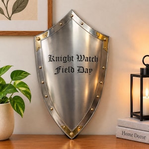 Pode incluir: Um escudo de metal prateado com ponta na parte superior e inferior, apresentando detalhes dourados nos cantos e rebites decorativos ao longo da borda. O centro do escudo exibe o texto preto Knight Watch Field Day em fonte gótica.
