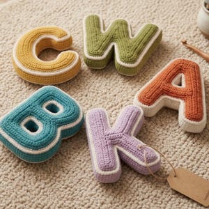 May include: Crocheted letters in various colors, including yellow, green, orange, teal, and purple, with white trim. The letters are arranged on a beige rug. A small succulent in a pot and a wooden crochet hook are also present.