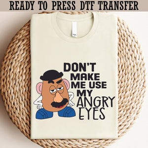 May include: A light beige t-shirt featuring a cartoon character with a black hat, mustache, and angry eyes. The text "DON'T MAKE ME USE MY ANGRY EYES" is printed in black. The shirt is displayed on a woven surface.