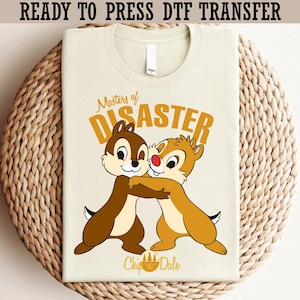 May include: A cream-colored t-shirt featuring Chip and Dale embracing, with the text "Masters of Disaster" in orange. The words "Chip & Dale" are below the characters. The phrase "READY TO PRESS DTF TRANSFER" is at the top.