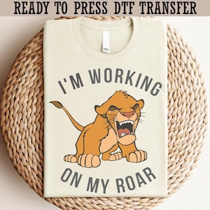 Może przedstawiać: Jasnobrązowy t-shirt z napisem "I'M WORKING ON MY ROAR" i ilustracją lwa z kreskówki. Lew jest pomarańczowy i brązowy, z otwartą paszczą. Koszulka jest złożona na tkanej powierzchni.