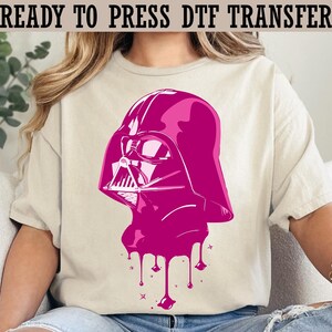 May include: A light beige t-shirt featuring a magenta graphic of Darth Vader's helmet with a dripping paint effect. The text "READY TO PRESS DTF TRANSFER" is printed at the top. The shirt is a casual style.