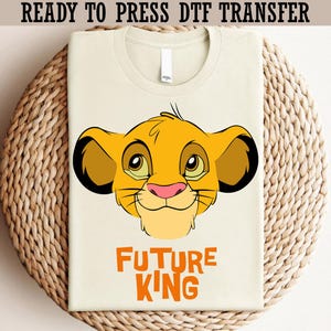 May include: Cream-colored t-shirt featuring a cartoon lion's face in yellow, brown, and pink, with the text "FUTURE KING" in orange. The shirt is folded and placed on a woven, circular surface. The text "READY TO PRESS DTF TRANSFER" is at the top.