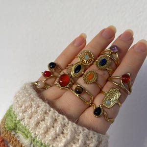 May include: A hand adorned with several gold rings, each featuring different coloured gemstones. The rings have various designs, including oval, round, and teardrop shapes. The hand is wearing a beige and multicoloured sweater, showcasing the rings.