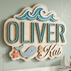 以下が含まれることがあります： 木製の壁掛けアート。名前「OLIVER」はティール色で、上部に波のデザインがあります。下にはクジラ、ヒトデ、タツノオトシゴ、名前「Kai」が表示されています。アートワークは、明るい緑色の壁に取り付けられています。