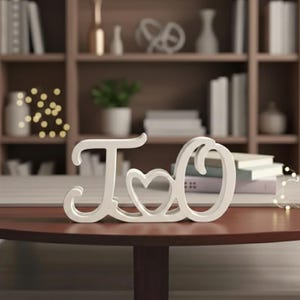Puede incluir: Un letrero decorativo blanco con las letras "I" y "O" y un símbolo de corazón. El letrero está sobre una mesa marrón oscuro. El fondo presenta una estantería con libros y adornos.