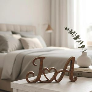 Puede incluir: Iniciales de madera "J" y "A" unidas por un corazón, sobre una mesa blanca. Un pequeño jarrón blanco con vegetación y una pila de libros también están presentes. El fondo muestra una cama con ropa de cama gris.
