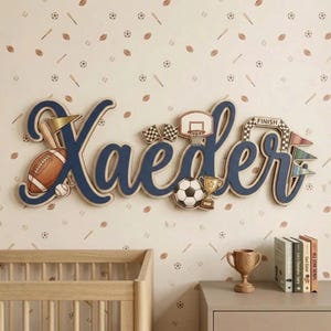 Op de afbeelding: Een gepersonaliseerd houten naambord voor aan de muur met de naam "Xaeder" in grote, donkerblauwe cursieve letters. Het ontwerp bevat sportthema-elementen zoals een football, een honkbal, een basketbalring, een gouden trofee en geblokte finishvlaggen.