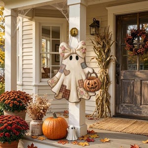 May include: A hanging ghost decoration featuring a large bow with floral patterns, patchwork fabric details, and a small jack-o'-lantern bucket. The ghost is cream-coloured with dark eyes and is suspended from a gold-coloured hook on a white porch post.