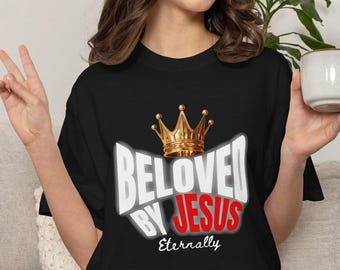May include: A black short-sleeved t-shirt featuring a graphic print with a gold crown above the white text "BELOVED BY" and the red text "JESUS" with the word "Eternally" in white script lettering positioned below the main graphic design.
