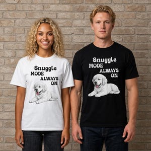 May include: A white t-shirt and a black t-shirt, each featuring a graphic of a golden retriever puppy lying down with its tongue out. Above the puppy, the text reads "SNUGGLE MODE ALWAYS ON" in a bold, rounded, dark-colored sans-serif font.