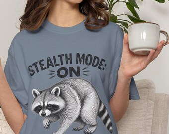 May include: A slate blue short-sleeve t-shirt featuring a black graphic of a raccoon and the text "STEALTH MODE: ON" printed in a distressed font. The raccoon is depicted in a realistic, detailed style with grey and white fur.