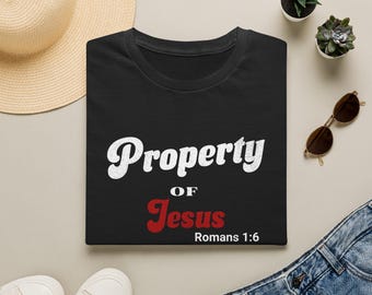 May include: A black t-shirt featuring the white text Property, the white text OF, and the red text Jesus, followed by the white text Romans 1:6. The shirt is displayed with a straw hat, two small potted succulents, sunglasses, and blue jeans.